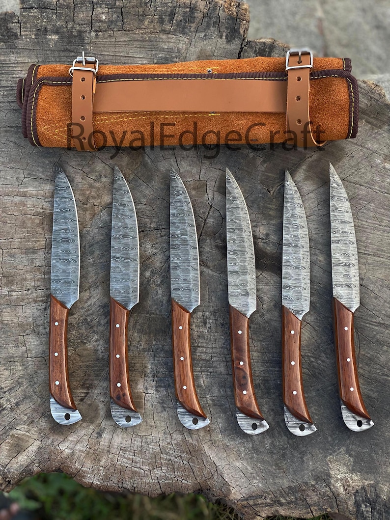 May include: A set of six steak knives with patterned blades and wooden handles, arranged on a wooden surface. The handles are a dark brown color with silver rivets. A brown leather knife roll with buckle closures is at the top. The text "RoyalEdgeCraft" is visible.