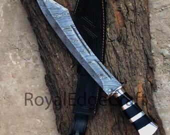 May include: A large, Damascus steel knife with a black and silver handle and a leather sheath. The blade has a wavy pattern. The handle features black and silver bands. The knife rests on a piece of wood.