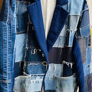 May include: A patchwork denim blazer in various shades of blue. The jacket features a contrasting dark blue collar and lapels, a single gold button closure, and multiple denim patches. The interior lining is white with a small yellow floral pattern.