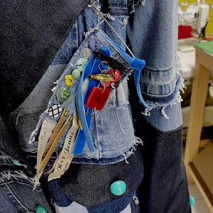 Pode incluir: Casaco de ganga patchwork com várias tonalidades de azul. O casaco apresenta botões decorativos, uma fita azul e uma coleção de pequenos amuletos e acessórios presos na lapela. As mangas têm uma borda desfiada.