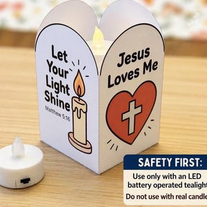 May include: A white paper lantern featuring a candle illustration with the text Let Your Light Shine Matthew 5:16 and a red heart with a cross and the text Jesus Loves Me. An LED battery operated tealight is placed next to the lantern.