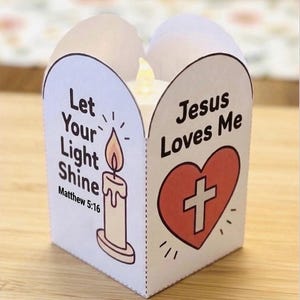 May include: A white paper candle wrap featuring a candle illustration with the text Let Your Light Shine Matthew 5:16 and a red heart with a white cross and the text Jesus Loves Me. A small LED candle is visible inside the folded paper structure.