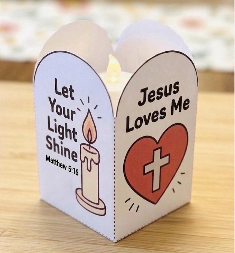 May include: A white paper candle wrap featuring a candle illustration with the text Let Your Light Shine Matthew 5:16 and a red heart with a white cross and the text Jesus Loves Me. A small LED candle is visible inside the folded paper structure.