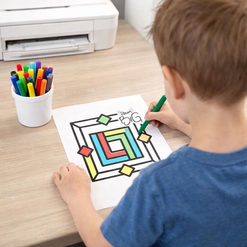 May include: A white sheet of paper with a black geometric line drawing and the text "Dream BIG" being coloured with a green marker. The drawing features nested squares and diamonds in red, blue, yellow, and green. A white cup holds various coloured markers.