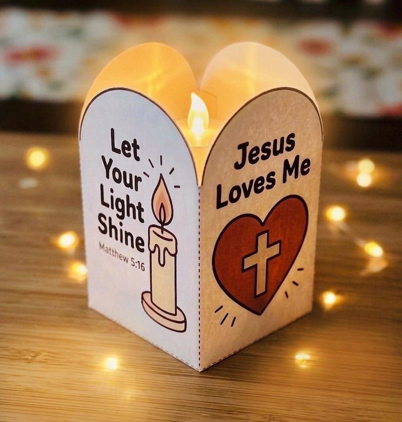 May include: A white paper lantern with a curved top edge featuring a lit LED candle inside. One side displays a candle illustration with the text Let Your Light Shine Matthew 5:16. The other side shows a red heart with a cross and Jesus Loves Me.