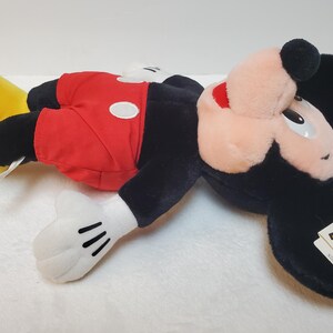May include: A plush Mickey Mouse toy, lying on its side. Mickey is wearing red shorts with white buttons, black shoes, and white gloves. His face is pink with black ears and a black nose. The toy has a tag attached.