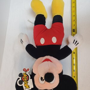 May include: A Mickey Mouse plush toy, upside down, with yellow feet, red shorts, and black body. The toy has white gloves and a tag that says "MOUSEKETOY'S". The toy measures approximately 46 cm in height.