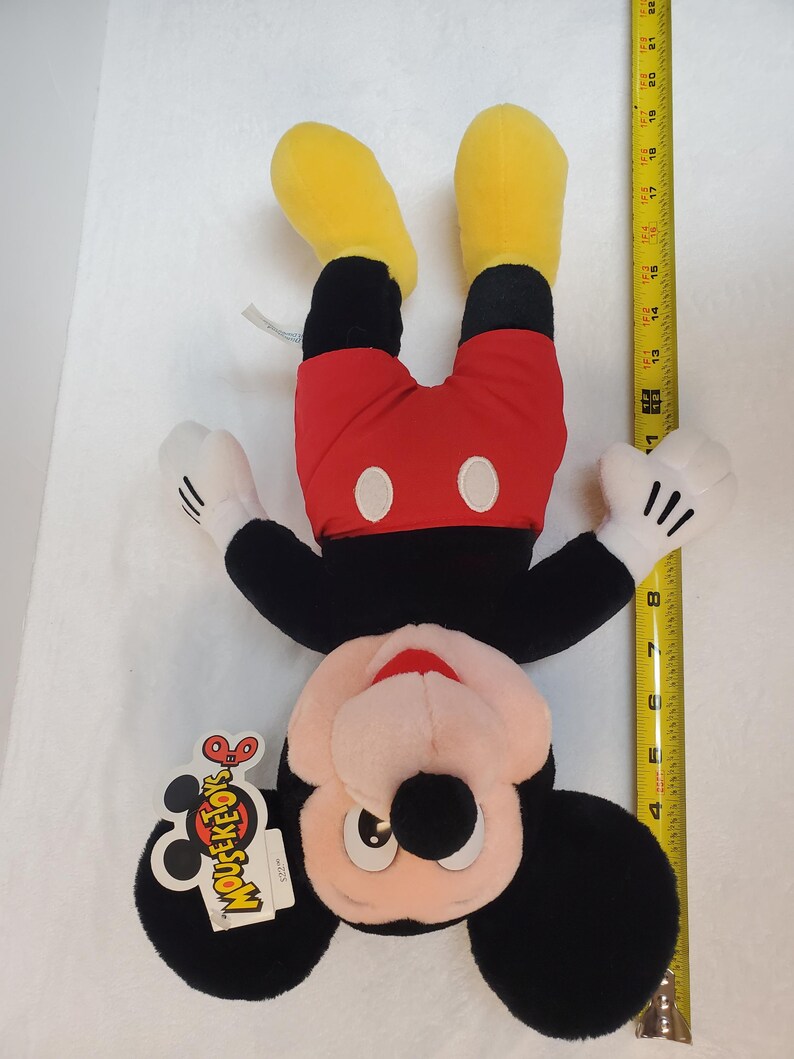 May include: A Mickey Mouse plush toy, upside down, with yellow feet, red shorts, and black body. The toy has white gloves and a tag that says "MOUSEKETOY'S". The toy measures approximately 46 cm in height.