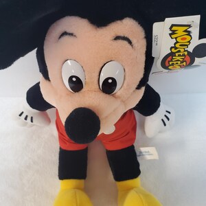 May include: A Mickey Mouse soft toy with large black ears, a pink face, and big white eyes. He wears red shorts, black arms and legs, and yellow shoes. A tag with the word "Mouseketeer" is attached.