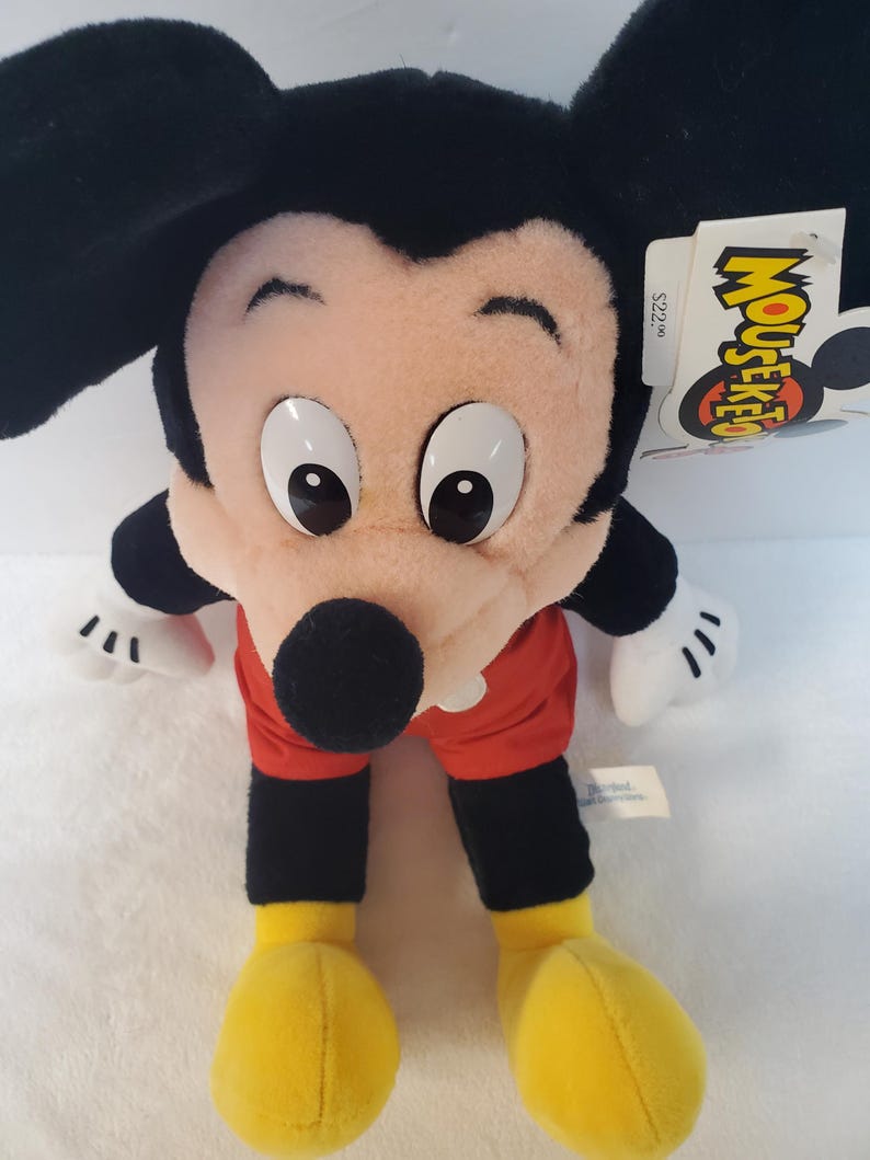 May include: A Mickey Mouse soft toy with large black ears, a pink face, and big white eyes. He wears red shorts, black arms and legs, and yellow shoes. A tag with the word "Mouseketeer" is attached.