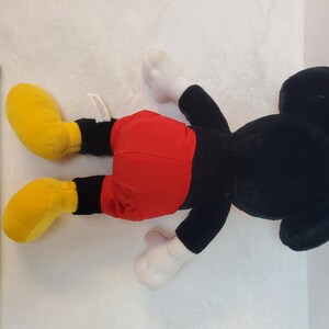 May include: A plush Mickey Mouse toy, lying on its back. The toy features a black body, red shorts, yellow shoes, and white gloves. A yellow tape measure is next to the toy, showing measurements in inches.
