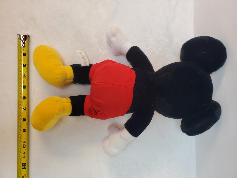 May include: A plush Mickey Mouse toy, lying on its back. The toy features a black body, red shorts, yellow shoes, and white gloves. A yellow tape measure is next to the toy, showing measurements in inches.