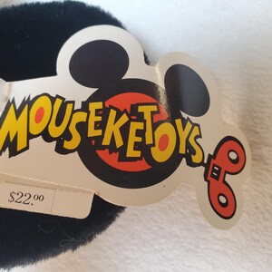 May include: A black plush toy with a white tag featuring the text "MOUSEKETOYS" in yellow and red. The tag has a Mickey Mouse head shape and a red and orange design. The tag also has a price tag.