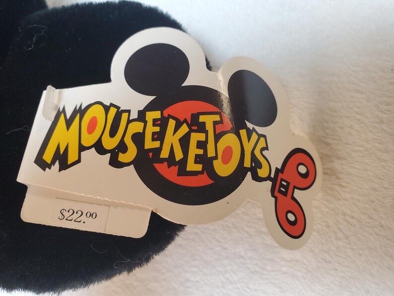 May include: A black plush toy with a white tag featuring the text "MOUSEKETOYS" in yellow and red. The tag has a Mickey Mouse head shape and a red and orange design. The tag also has a price tag.