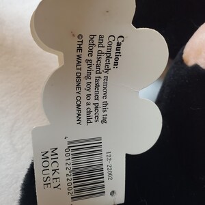 May include: A black Mickey Mouse plush toy with a white tag. The tag has the text "MICKEY MOUSE" and a barcode. The tag also includes a caution about removing fasteners before giving the toy to a child.