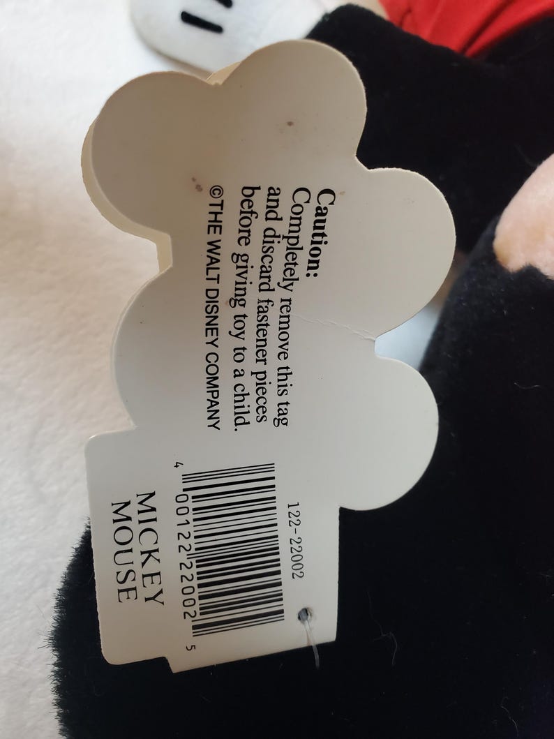 May include: A black Mickey Mouse plush toy with a white tag. The tag has the text "MICKEY MOUSE" and a barcode. The tag also includes a caution about removing fasteners before giving the toy to a child.