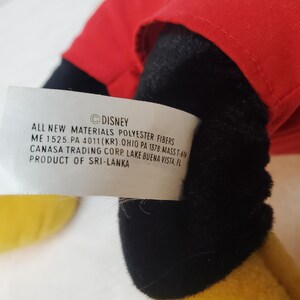 May include: A close-up of a plush toy with a red top, black shorts, and yellow shoes. A white tag with the Disney logo and product information is visible.