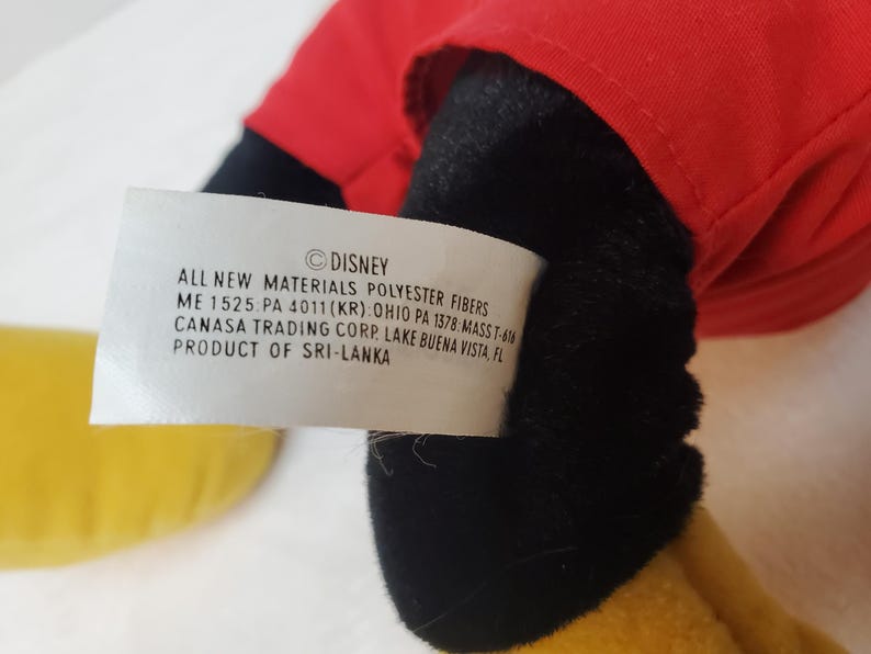 May include: A close-up of a plush toy with a red top, black shorts, and yellow shoes. A white tag with the Disney logo and product information is visible.