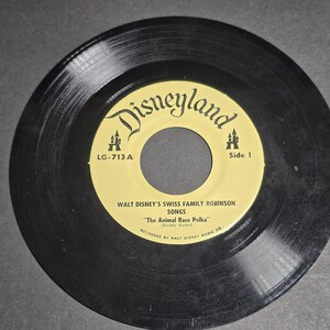 Op de afbeelding: Een vintage 7-inch vinylplaat met een geel en zwart label met het Disneyland-logo. Op het label staat "Walt Disney's Swiss Family Robinson Songs" en "The Animal Race Polka". De plaat is een verzamelobject.