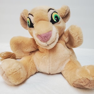 May include: A plush toy lioness with a light brown body, a pink nose, and green and black eyes. The toy has a friendly expression and is lying down with its paws outstretched. The toy is made of soft, fuzzy material.