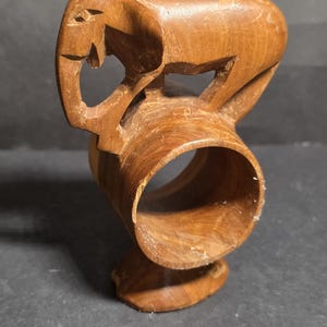 May include: A carved wooden napkin ring featuring an elephant design. The ring is made of light brown wood and has a circular opening. The elephant is carved in a stylized manner, with a trunk and ears visible. The napkin ring is a decorative item.