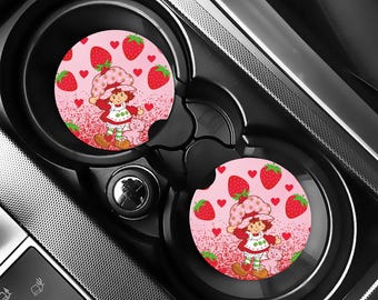 Könnte beinhalten: Zwei Auto-Untersetzer mit einer Cartoonfigur mit Erdbeerhut, Kleid und Stiefeln, umgeben von Erdbeeren und Herzen auf rosa Hintergrund. Die Untersetzer befinden sich in den Getränkehaltern eines Autos.