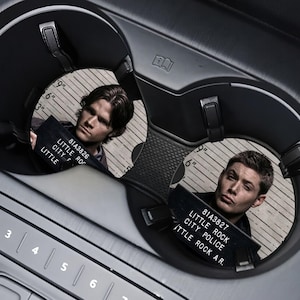 May include: Two car coasters with images of men holding mugshot signs. The signs read "LITTLE ROCK CITY POLICE" and include the numbers "81A3826" and "81A3827". The coasters are set in a black car console.