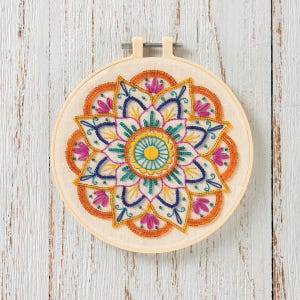May include: Embroidered mandala art in a wooden hoop. The design features a symmetrical pattern with orange, blue, yellow, teal, and pink thread on a cream-colored fabric. The hoop is held by a metal clasp, creating a finished piece.