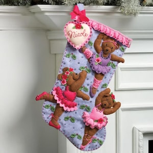 Peut inclure: Une chaussette de Noël avec un fond bleu clair, décorée de trois ours en peluche bruns vêtus de tenues de ballet roses et violettes. La chaussette a une bordure rose et une étiquette en forme de cœur avec le mot "Nicole".