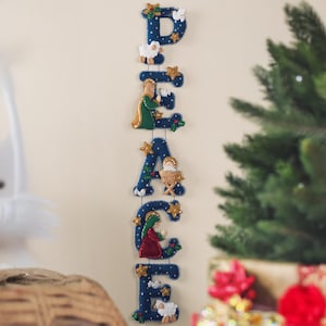 Puede incluir: Decoración navideña de fieltro azul que deletrea "PEACE" verticalmente. Cada letra está adornada con elementos festivos como estrellas, ovejas y figuras que representan la escena de la Natividad. La decoración está colgada en una pared, con un árbol de Navidad visible en el fondo.