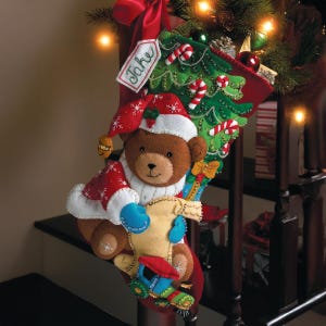 May include: A festive Christmas stocking featuring a teddy bear in a Santa outfit. The red stocking has a green Christmas tree design and candy cane decorations. A tag with the name "Jake" is visible.