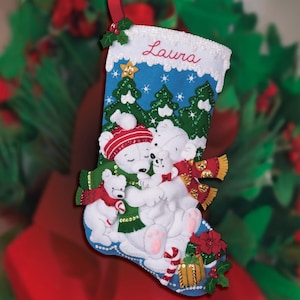 Op de afbeelding: Een blauwe kerstsok met een witte manchet, met een familie van witte ijsberen met rode en groene sjaals en mutsen. De sok is versierd met groene bomen, sneeuwvlokken en de naam "Laura" in het rood.
