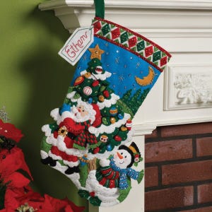 Peut inclure: Une chaussette de Noël au design festif. La chaussette représente le Père Noël, un bonhomme de neige et un sapin de Noël décoré sur fond de ciel bleu. Le haut de la chaussette présente un motif rouge, vert et blanc. Une étiquette avec le nom "Ethan" est attachée.