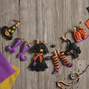 May include: A Halloween-themed felt garland featuring a black witch hat, purple boots, a black dress with an orange sash, striped stockings, and a black cat, all suspended from a brown twine string with small wooden clothespins.