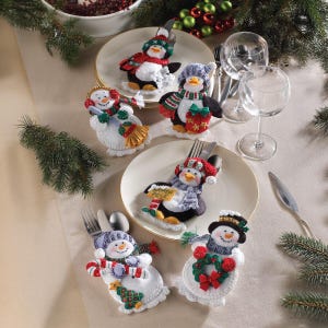 Puede incluir: Una mesa festiva navideña con portacubiertos de fieltro con forma de muñecos de nieve y pingüinos. Cada soporte tiene un diseño único con bufandas, sombreros y accesorios coloridos. Los soportes están sobre platos blancos.