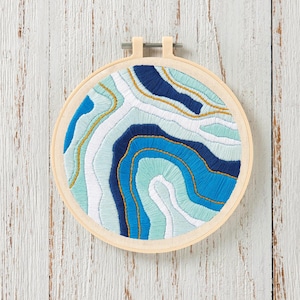 Peut inclure: Un cercle à broder avec un motif abstrait dans les tons de bleu, blanc et sarcelle, avec des accents de fil doré. Le motif ressemble à une carte topographique ou à une tranche d'agate. Le cercle est en bois clair.