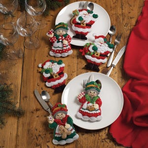 May include: A festive holiday table setting featuring white plates with felt Santa Claus and Mrs. Claus figures. The figures are red, white, and green, with details like belts and hats. Silverware and wine glasses are also visible.