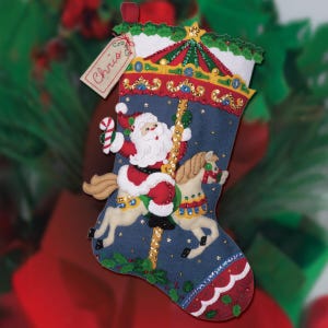 Può includere: Una calza di Natale a tema giostra. Babbo Natale cavalca un cavallo della giostra, con un bastoncino di zucchero in mano. La calza è blu con dettagli dorati e rossi, e il nome "Chris" è su un'etichetta.