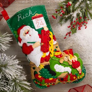 Puede incluir: Un calcetín navideño de fieltro verde con un diseño de Papá Noel. El calcetín presenta el nombre "Rick" y un calendario que muestra el 24 de diciembre. Papá Noel sostiene una lista. La parte inferior del calcetín tiene un tren, un oso de peluche y una bailarina.