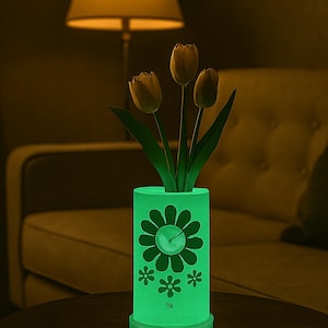 Puede incluir: Un jarrón cilíndrico verde brillante con un diseño floral, que contiene tres tulipanes amarillos. El jarrón está iluminado, proyectando una luz suave, y está sobre una mesa oscura. Una lámpara y un sofá están en el fondo.