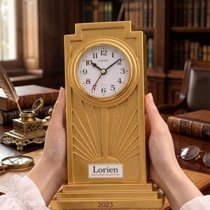 Puede incluir: Un reloj dorado con esfera redonda y números romanos. El reloj tiene una base rectangular y un diseño decorativo de rayos de sol. La esfera del reloj indica "Lorien" y "2023".