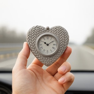 Könnte beinhalten: Eine graue herzförmige Uhr mit strukturierter Oberfläche, mit einem runden Zifferblatt mit römischen Ziffern. Die Zeiger der Uhr zeigen die Uhrzeit an. Die Uhr wird in einer Hand gehalten.