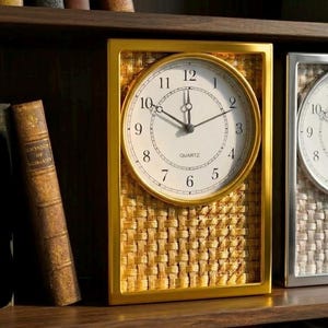 May include: Two quartz clocks with white faces and black hands sit on a wooden shelf. One clock has a gold frame, the other a silver frame. Both have woven backgrounds. Antique books line the shelf.