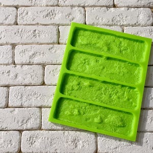 May include: A bright lime green silicone mold featuring four rectangular cavities with a textured, uneven surface pattern designed to replicate the appearance of natural stone or brick masonry for casting decorative elements.
