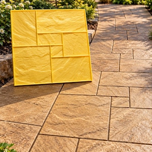 May include: A square yellow concrete stamp mold featuring a multi-rectangular pattern with a textured stone-like surface. The mold is positioned upright on a finished walkway that displays the same stone pattern in a tan color.