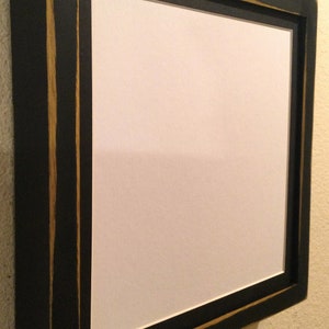 May include: A black wooden picture frame with a white interior. The frame has a distressed finish with gold accents and decorative metal studs. The frame is mounted on a beige wall.