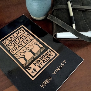 May include: A black book titled "Psalms Poems & Prayers" with a tan and black graphic of trees. A leather-bound journal with a pen and a small blue ceramic vase are also on the wooden surface.