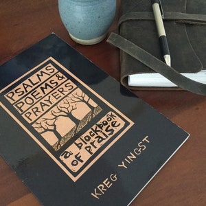May include: A black book with the title "Psalms Poems & Prayers a blockbook of praise" by Kreg Yingst. The book is on a wooden table with a blue ceramic mug and a brown leather journal with a pen.