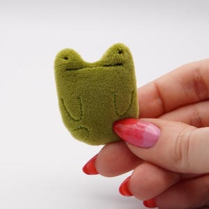 May include: A small, olive green frog-shaped plushie with a stitched mouth and eyes. The plushie is held in a hand with red and pink painted nails against a white background. The frog has a simple, minimalist design.