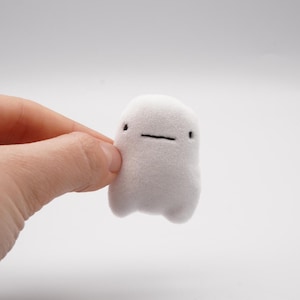 May include: A small, white plush toy with a minimalist design. The toy has a rounded body, small black eyes, and a horizontal black line for a mouth. It is held by a hand against a plain white background.